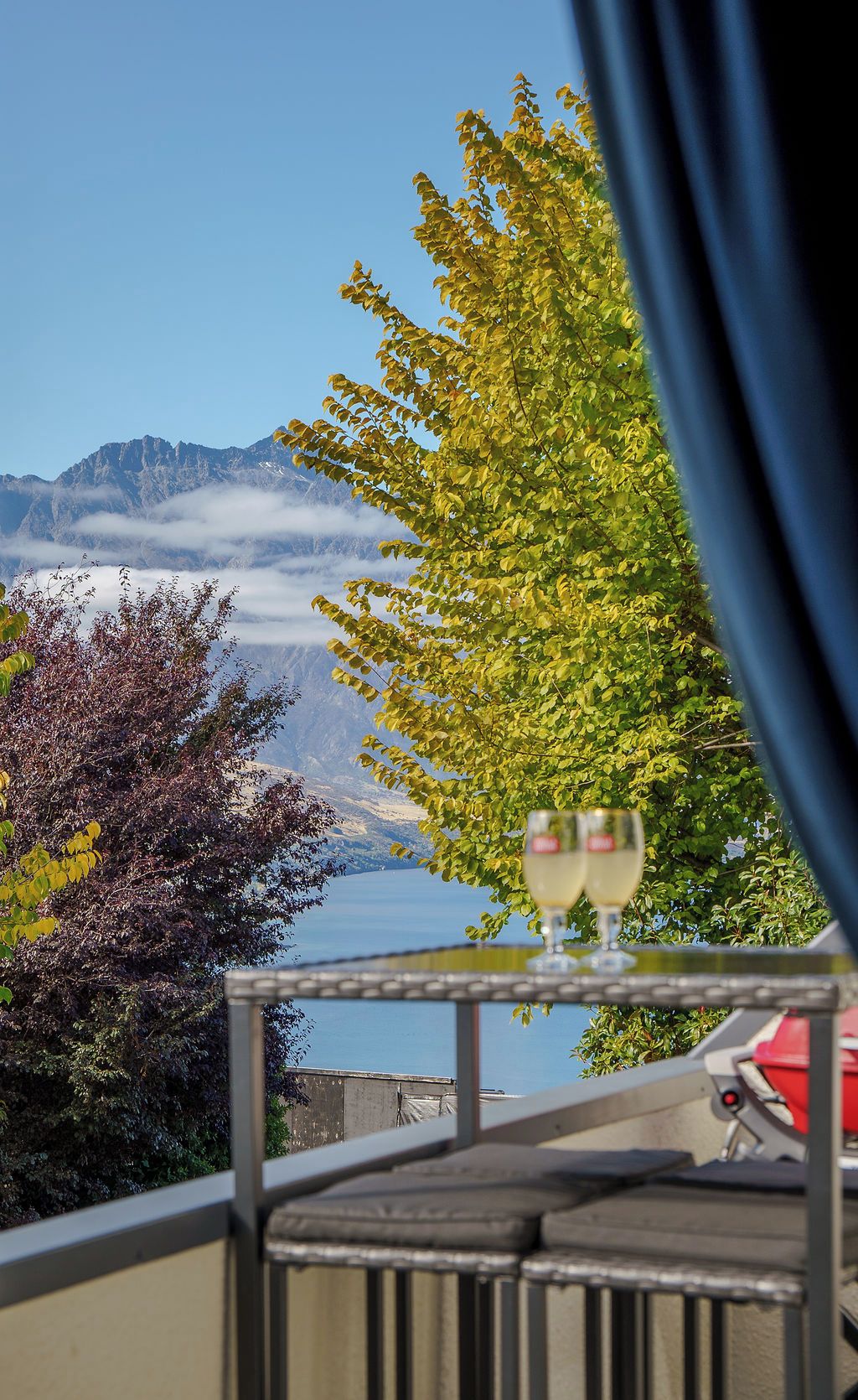A balcony with a table, two wine glasses, and a view of a mountain range and lake.