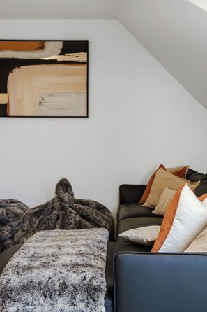 A modern bedroom with a sloped ceiling, a large abstract painting, a dark sofa with multiple cushions, and a fur throw. - Sarahs Scenic Stay | Mountain Lake Views (Queenstown)