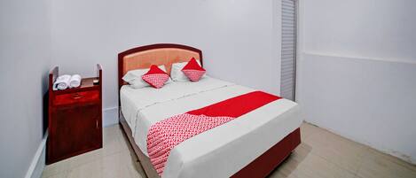 Una cama bien hecha con una colcha roja y blanca, una mesita de noche de madera con toallas y un armario cerrado.
