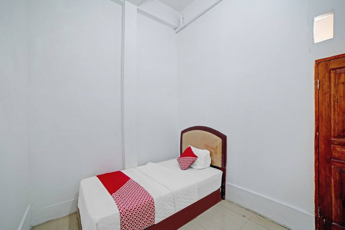 Un llit petit i ben fet amb un capçal de fusta a una habitació de parets blanques i una porta de fusta.