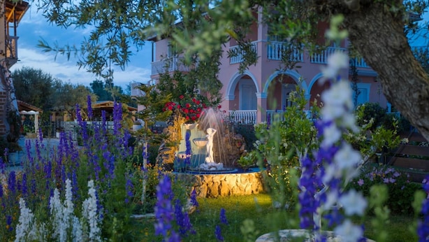 A garden with a fountain, surrounded by various flowers and a building with balconies in the background. - Antigoni s Family House - Antigoni s Family House (Zakynthos)