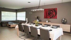 A modern dining area with a long table set for six, surrounded by chairs. There is a sofa and ottoman in the background, a large window with blinds, and a wall-mounted rose painting. - Casa Tibella is an attractively furnished holiday home. (Ichtegem)