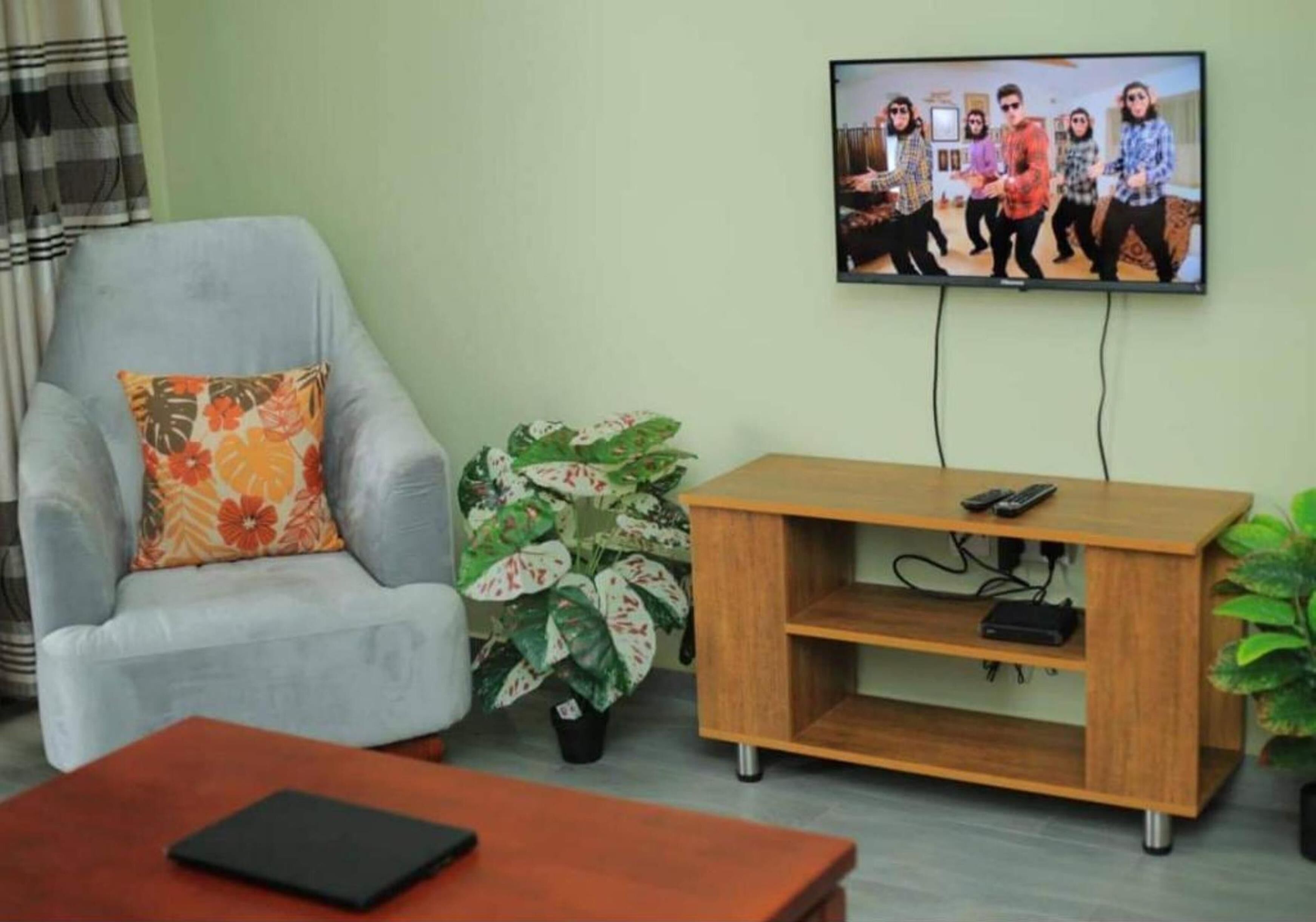 A living room with a grey armchair, a wooden coffee table, a TV mounted on the wall, and a potted plant.