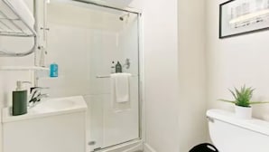 Ein modernes Badezimmer mit einer Duschkabine aus Glas, einem weißen Waschbecken und einer Toilette. An der Wand hängt ein gerahmtes Bild und auf dem Toilettenbecken steht ein kleiner Blumentopf.