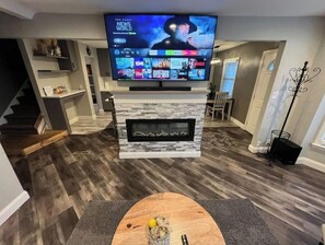 Una sala moderna con un televisor de pantalla plana sobre la chimenea, un comedor con mesa de madera, y una escalera que sube a otro nivel.