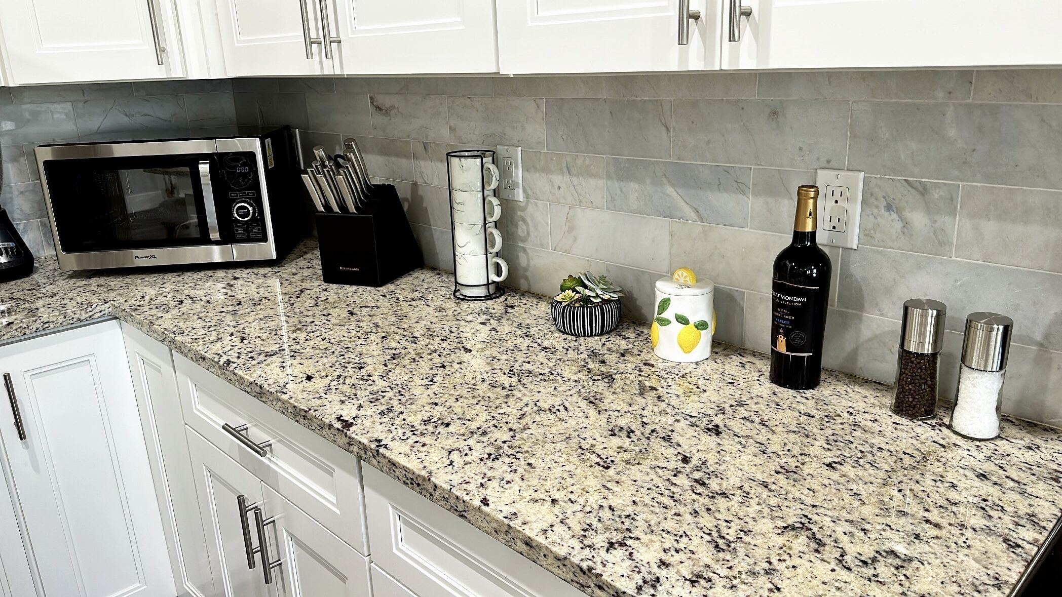 A kitchen with white cabinets, a granite countertop, a microwave, a coffee maker, a wine bottle, and salt and pepper shakers.