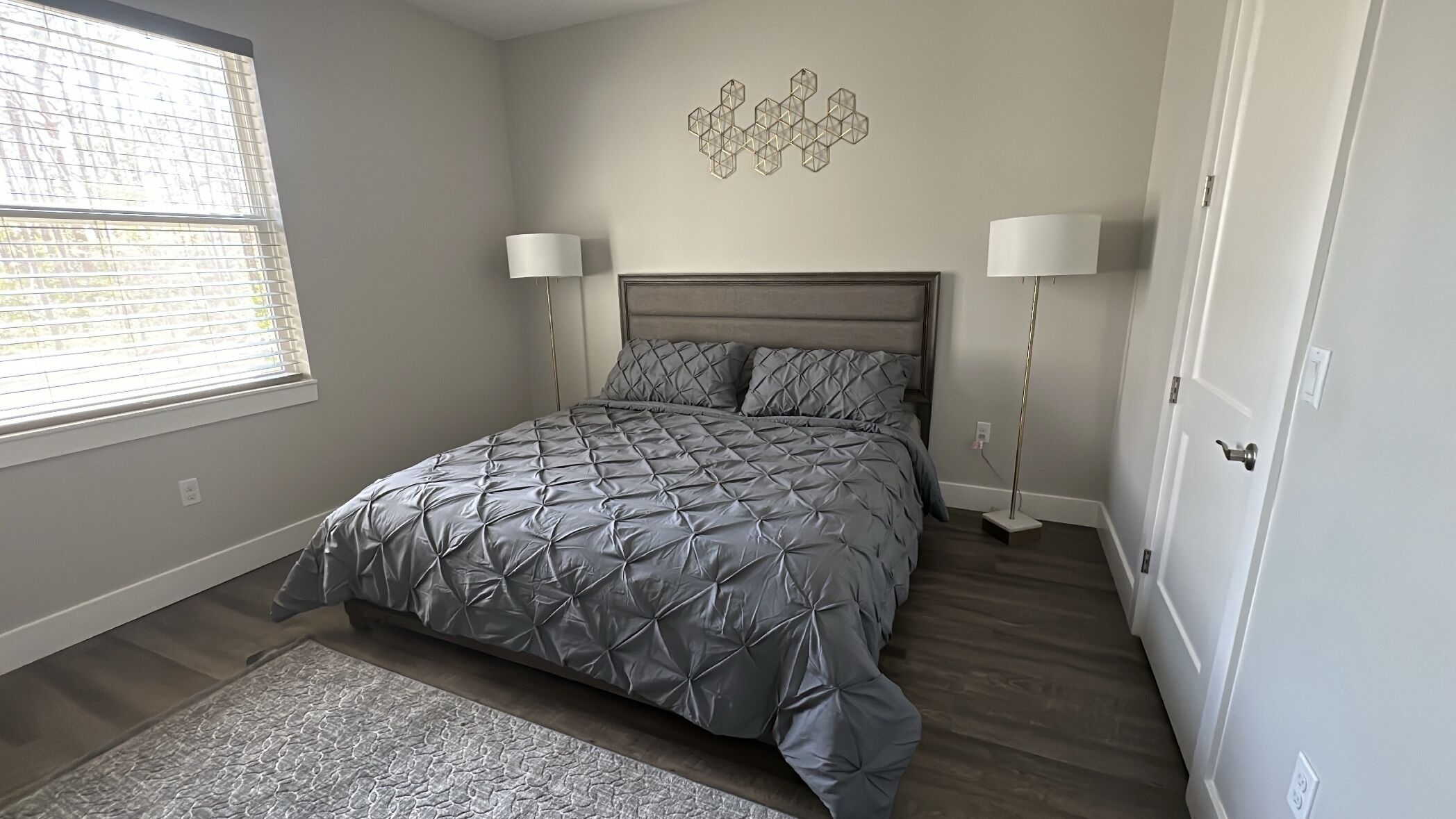 A bedroom with a bed, two bedside lamps, a wall art piece, and a window with blinds.