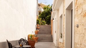 Un estrecho callejón con suelo de hormigón, flanqueado por paredes blancas y de piedra, que conduce a una escalera. Hay mesas y sillas al aire libre, y varias plantas en macetas a lo largo del camino.