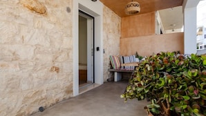 Un pasillo de paredes de piedra con una puerta que da a un dormitorio, un banco con cojines y una planta grande en maceta.