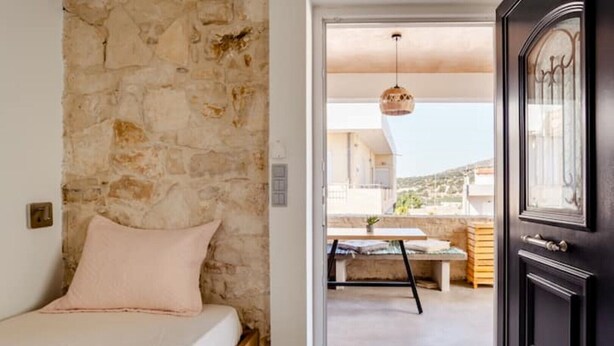 Un dormitorio con pared de piedra, una cama con almohada rosa y vista a un balcón exterior con mesa y lámpara colgante.