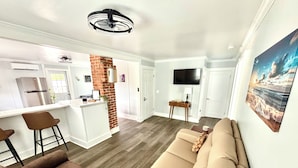 Un salon moderne avec un canapé beige, un fauteuil marron et un coin cuisine agrémenté d’un mur en briques apparentes.