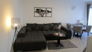 Ein modernes Wohnzimmer mit einer Eckcouch, einem kleinen runden Tisch und einem Essbereich mit Stühlen. An der Wand befinden sich gerahmte Kunstwerke und eine Uhr.