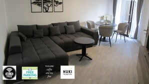 Ein modernes Wohnzimmer mit einem Ecksofa, einem kleinen runden Tisch und einem Essbereich mit Stühlen. An der Wand befinden sich gerahmte Kunstwerke, eine Uhr und ein Fenster mit Vorhängen.