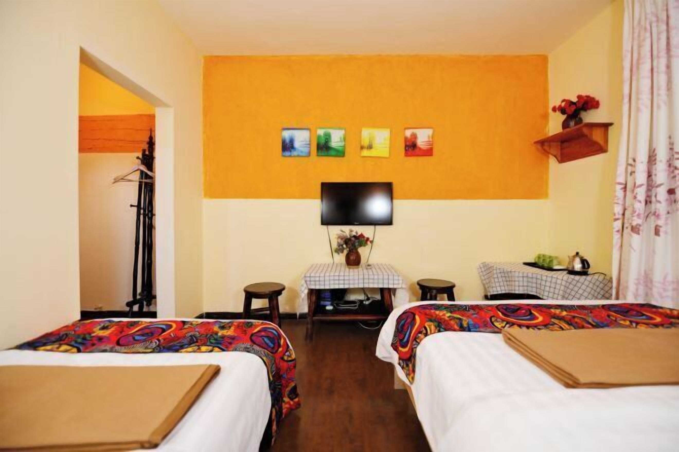 A hotel room with two beds, a television, a small table with a vase, and a wall with four framed pictures.