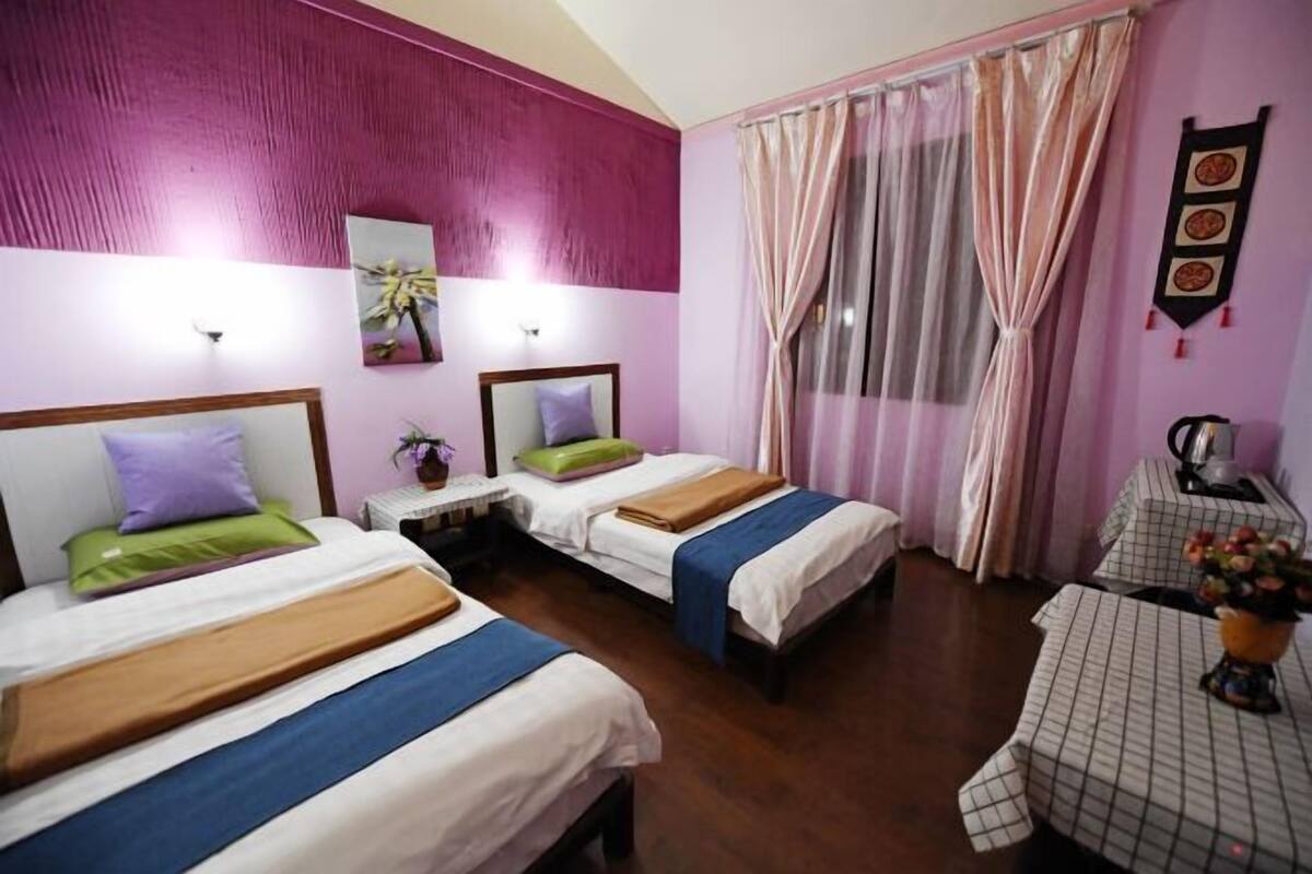 A hotel room with two beds, a small table with a checkered tablecloth, and a window with sheer curtains.