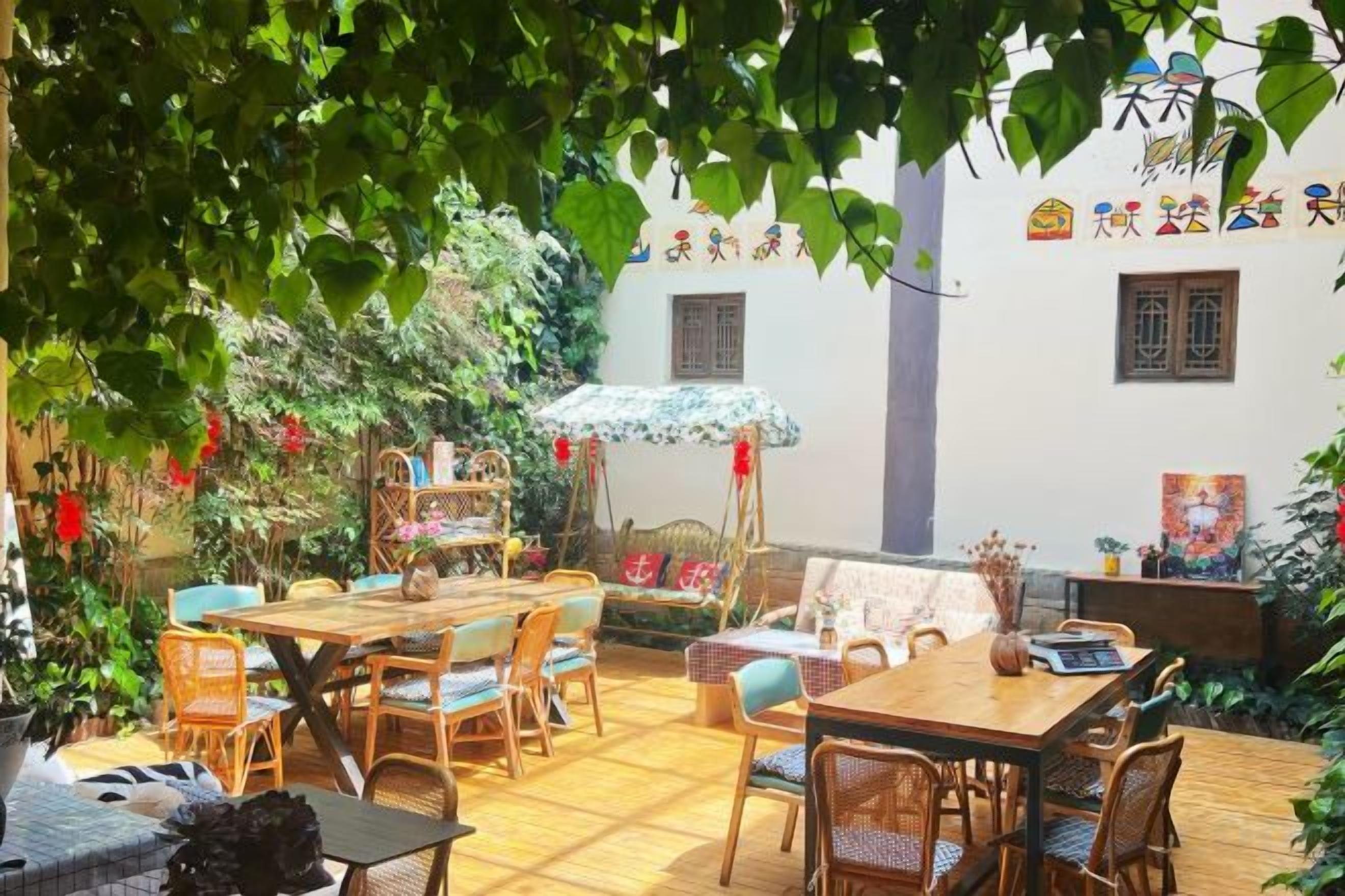 An outdoor patio with wooden tables and chairs, surrounded by greenery and a white building with traditional Chinese decorations.