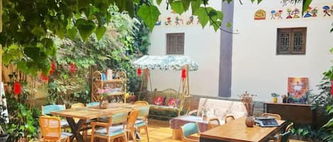 An outdoor patio with wooden tables and chairs, surrounded by greenery and a white building with traditional Chinese decorations.