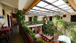 Amplio espacio interior con techo alto, vigas de madera y diversas plantas. Hay varias zonas de estar y una escalera que lleva a un nivel superior.