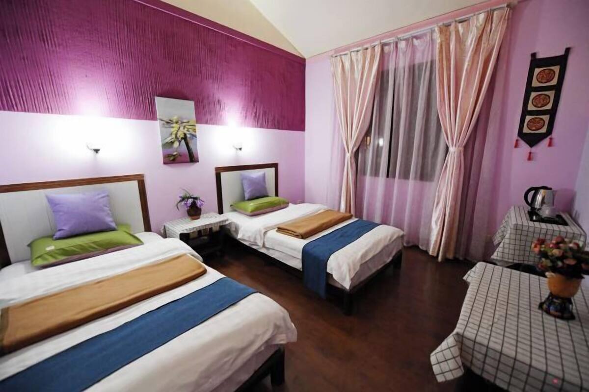 A hotel room with two beds, a small table with a vase of flowers, and a window with sheer curtains.