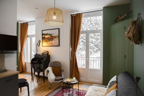A cozy living room with a sofa, a dining table, and a TV. There is a window with curtains, a painting on the wall, and a coat hanging on the wall.