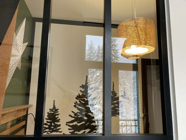 A modern interior with a large window reflecting snowy trees, a hanging woven light fixture, and a wooden staircase.