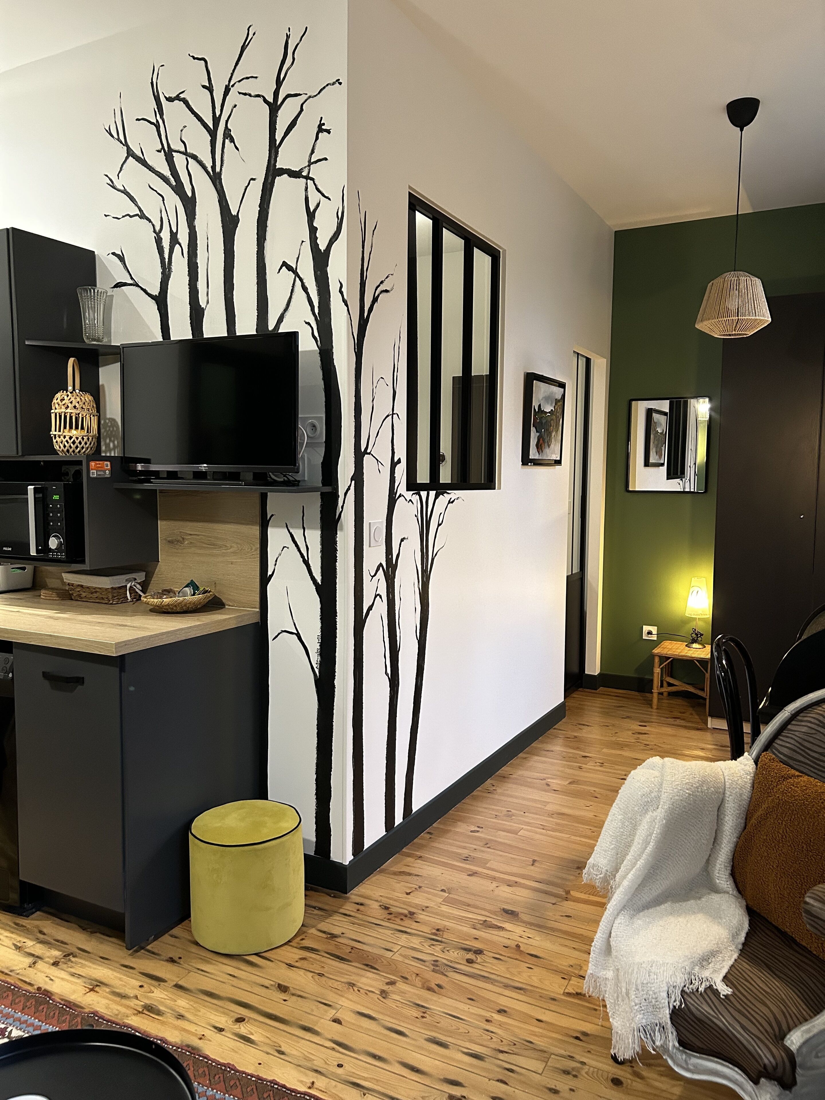 A modern living space with a kitchen area featuring a microwave, a coffee machine, and a small table with a basket. There is a yellow ottoman, a white throw blanket on a brown sofa, and a wooden floor.