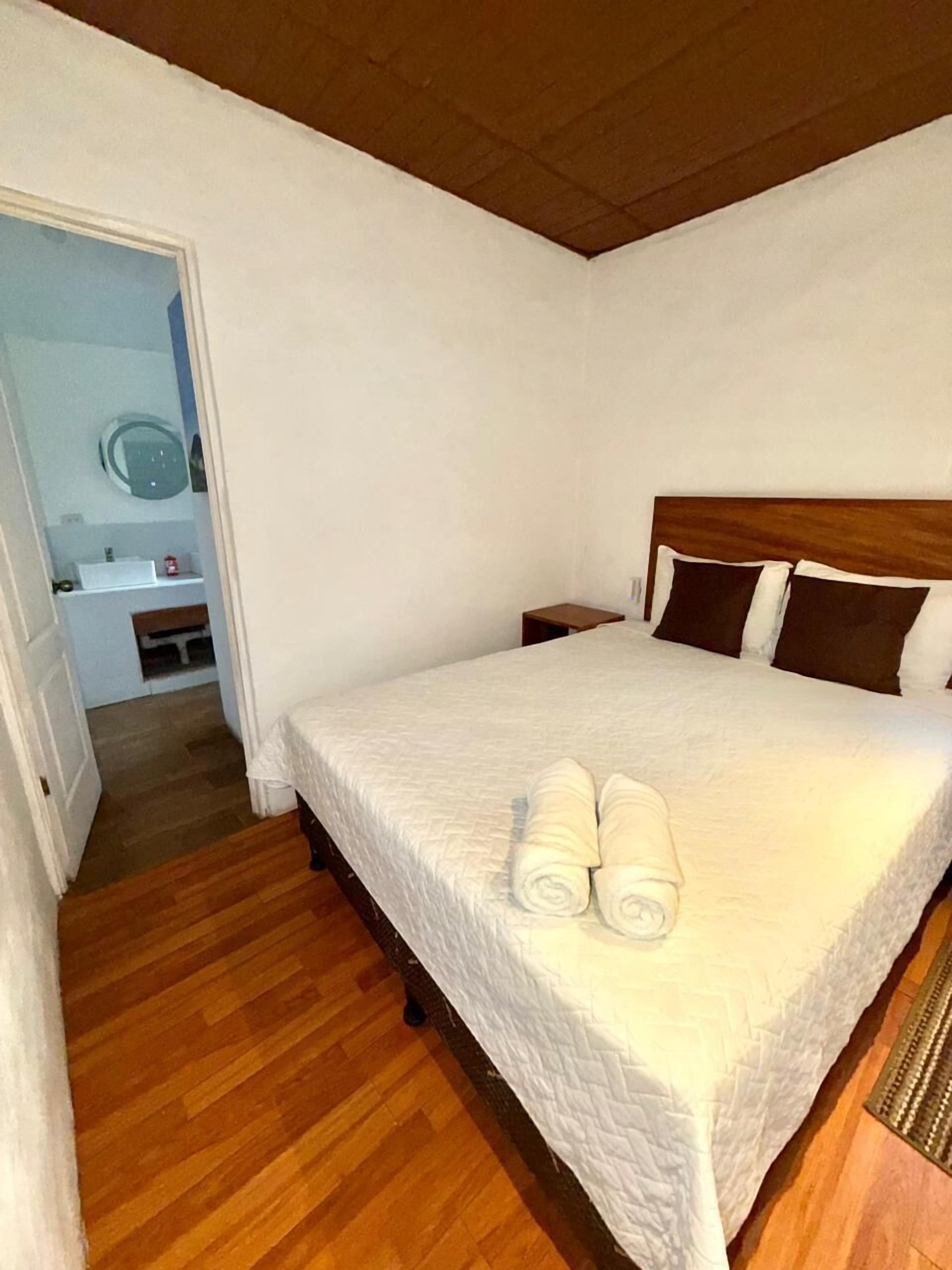 Een slaapkamer met een houten vloer, een bed met een witte sprei, twee opgerolde handdoeken, een houten hoofdbord en een nachtkastje.