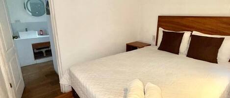 A bedroom with a wooden floor, a bed with a white quilt, two rolled towels, a wooden headboard, and a nightstand.