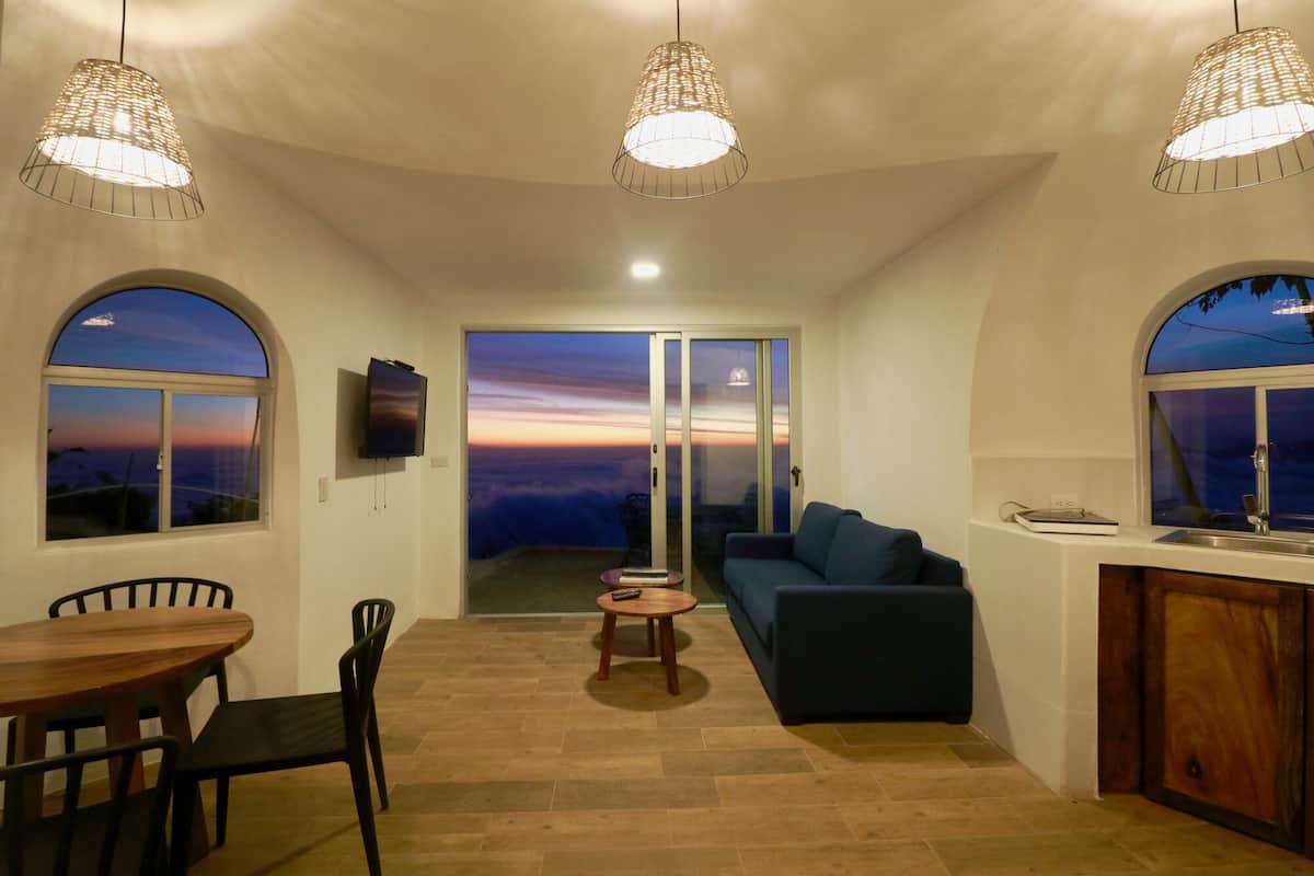 A cozy living room with a sofa, a dining table with chairs, and a TV mounted on the wall. There are large windows with a view of the sunset.