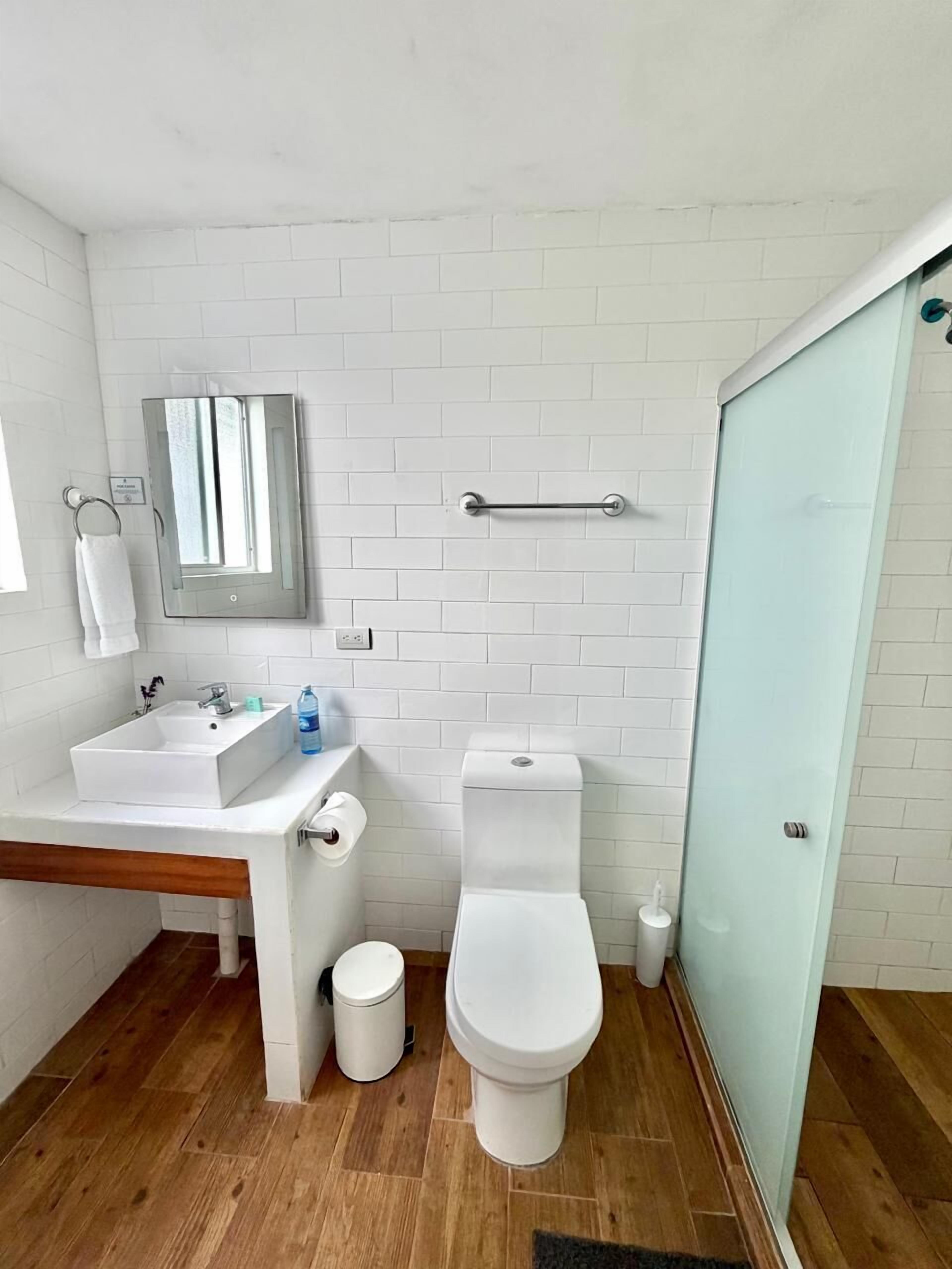 A small, modern bathroom with white tiles, a wooden floor, a white sink, a toilet, and a glass shower enclosure.