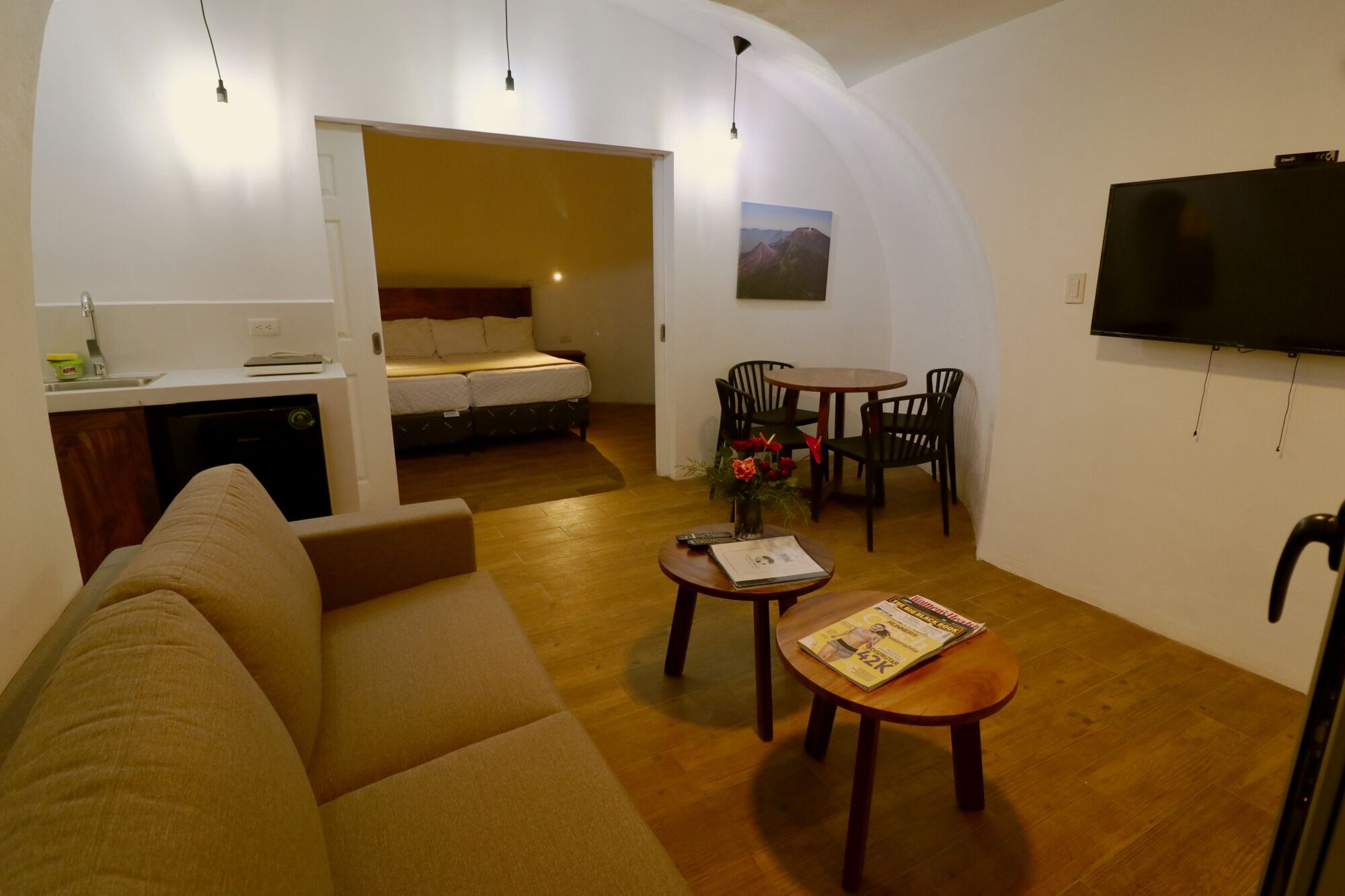 A compact living space with a sofa, a dining table with chairs, a small kitchen area, and a TV mounted on the wall.