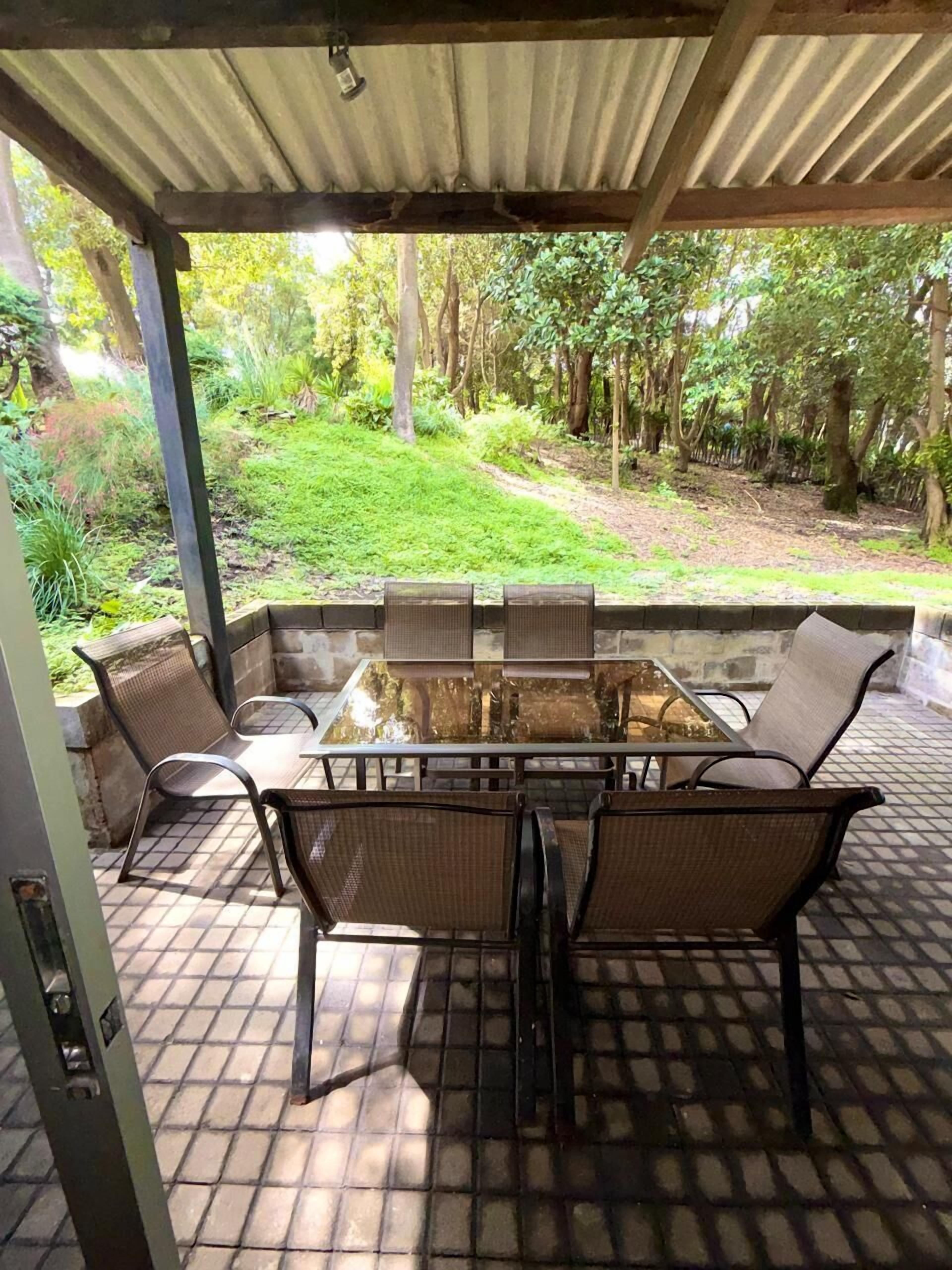 An outdoor patio with a glass-top table and wicker chairs, surrounded by a tiled floor and a metal roof.