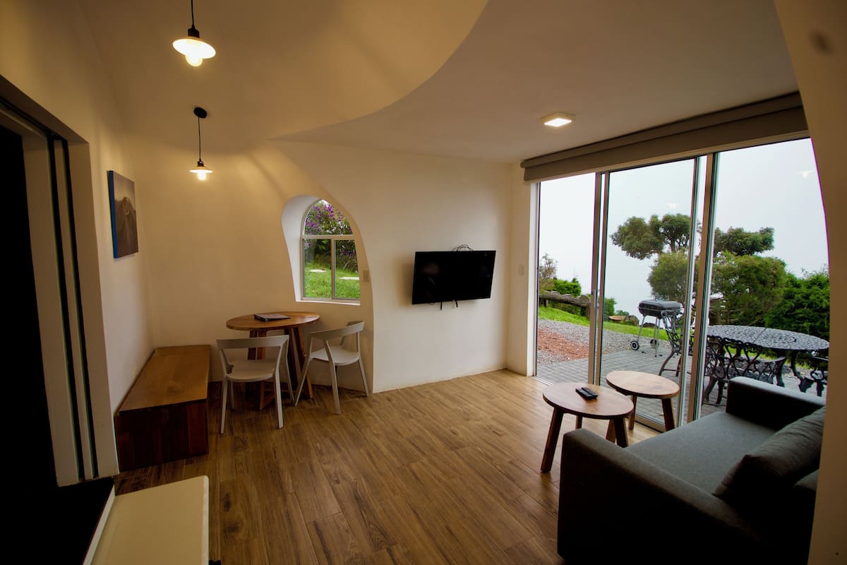A modern living room with a wooden floor, a sofa, a TV mounted on the wall, and a large window offering a view of the outdoors.