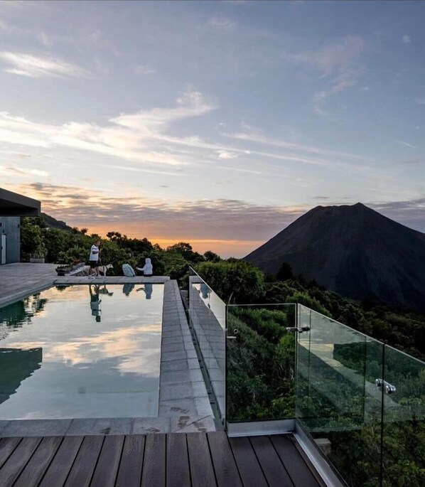 Piscina moderna al aire libre con barandillas de cristal, rodeada de exuberante vegetación y vistas a la montaña.