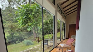 A room with large windows, wooden flooring, wicker furniture, and a view of a garden. - Casa Mia Suite with Jacuzzi and Panoramic View (Monteverde)
