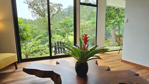 A room with a large wooden table, a potted plant, and a magazine. There are large windows with a view of greenery outside. - Casa Mia Suite with Jacuzzi and Panoramic View (Monteverde)