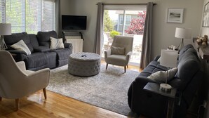 Ein modernes Wohnzimmer mit einer grauen Sofagarnitur, einem runden Hocker, einem Couchtisch und einem an der Wand montierten Fernseher.