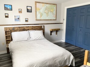 Quarto com cabeceira de madeira, cama com roupa de cama branca, mapa-múndi na parede e porta azul.