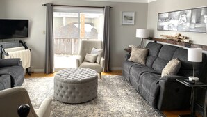 Ein Wohnzimmer mit grauem Sofa, Hocker und Sesseln, einem Flachbildfernseher und einem großen Fenster mit Vorhängen.
