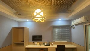 A compact hotel room with a desk, a television, a window with blinds, a ceiling light fixture, and an air conditioning unit. - Beicheng Hotel (Dazhu)