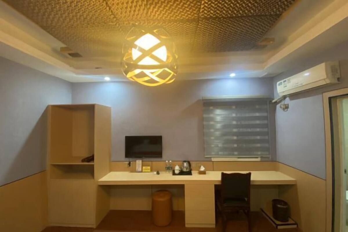 A compact hotel room with a desk, a television, a window with blinds, a ceiling light fixture, and an air conditioning unit.