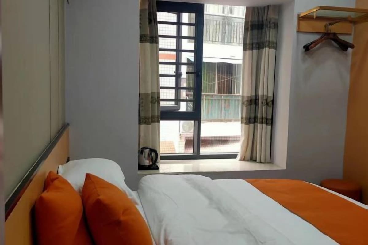 Une chambre d’hôtel avec un lit, une fenêtre avec des rideaux, un porte-manteau et une petite table avec une bouilloire.