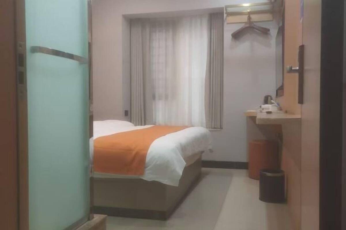 A hotel room with a bed, a bedside table, a mirror, a hanging coat rack, and a trash bin.