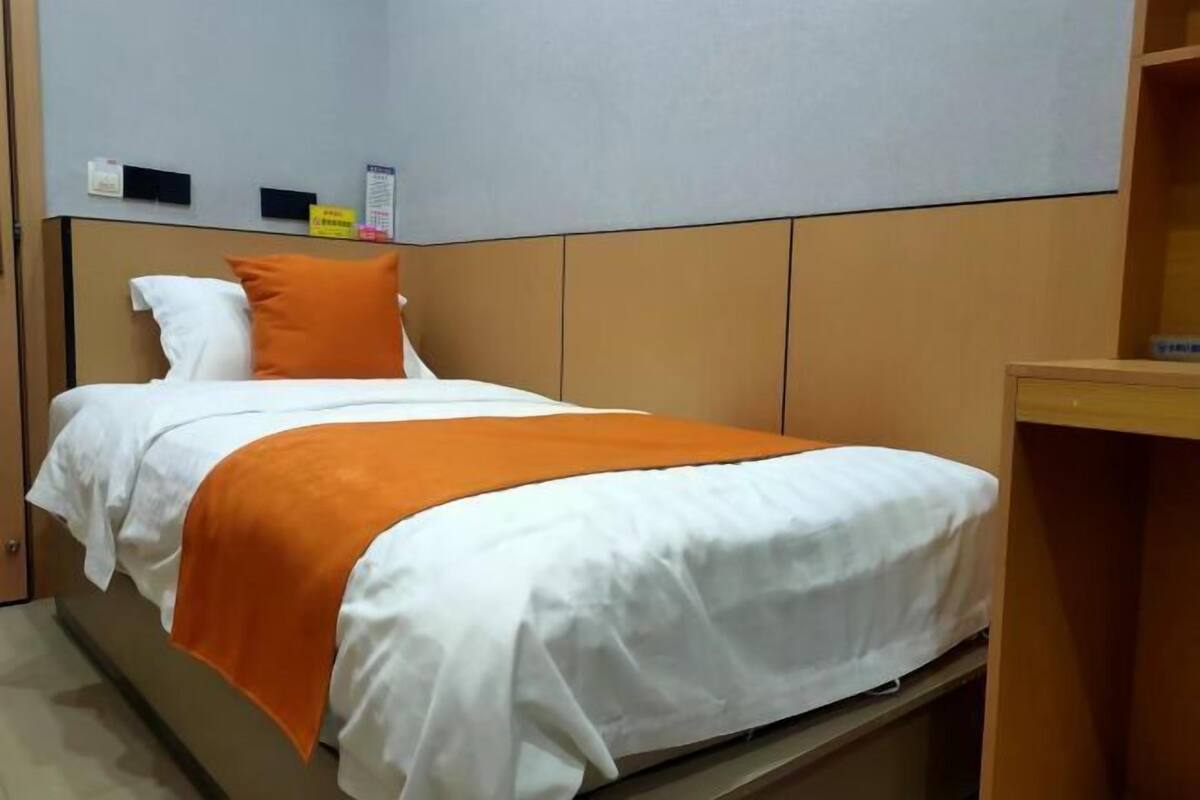 A single bed with white and orange bedding, a wooden headboard, and a small shelf with items on the wall above.