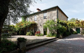 A stone building with a green door, surrounded by lush vegetation and a cobblestone pathway. - Casa de Abbades (Ponte de Lima)