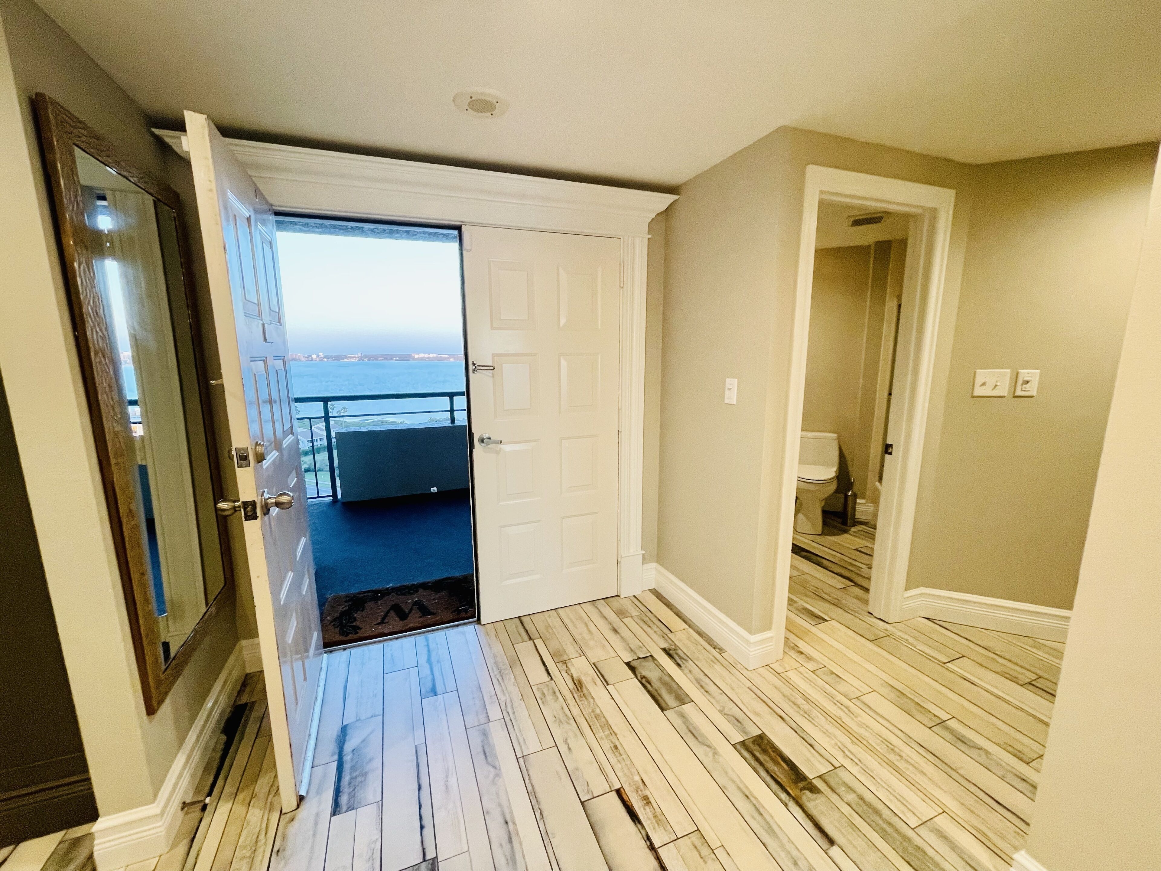 A hallway with a wooden floor, a white door leading to a balcony with a view, and a bathroom visible through an adjacent door.