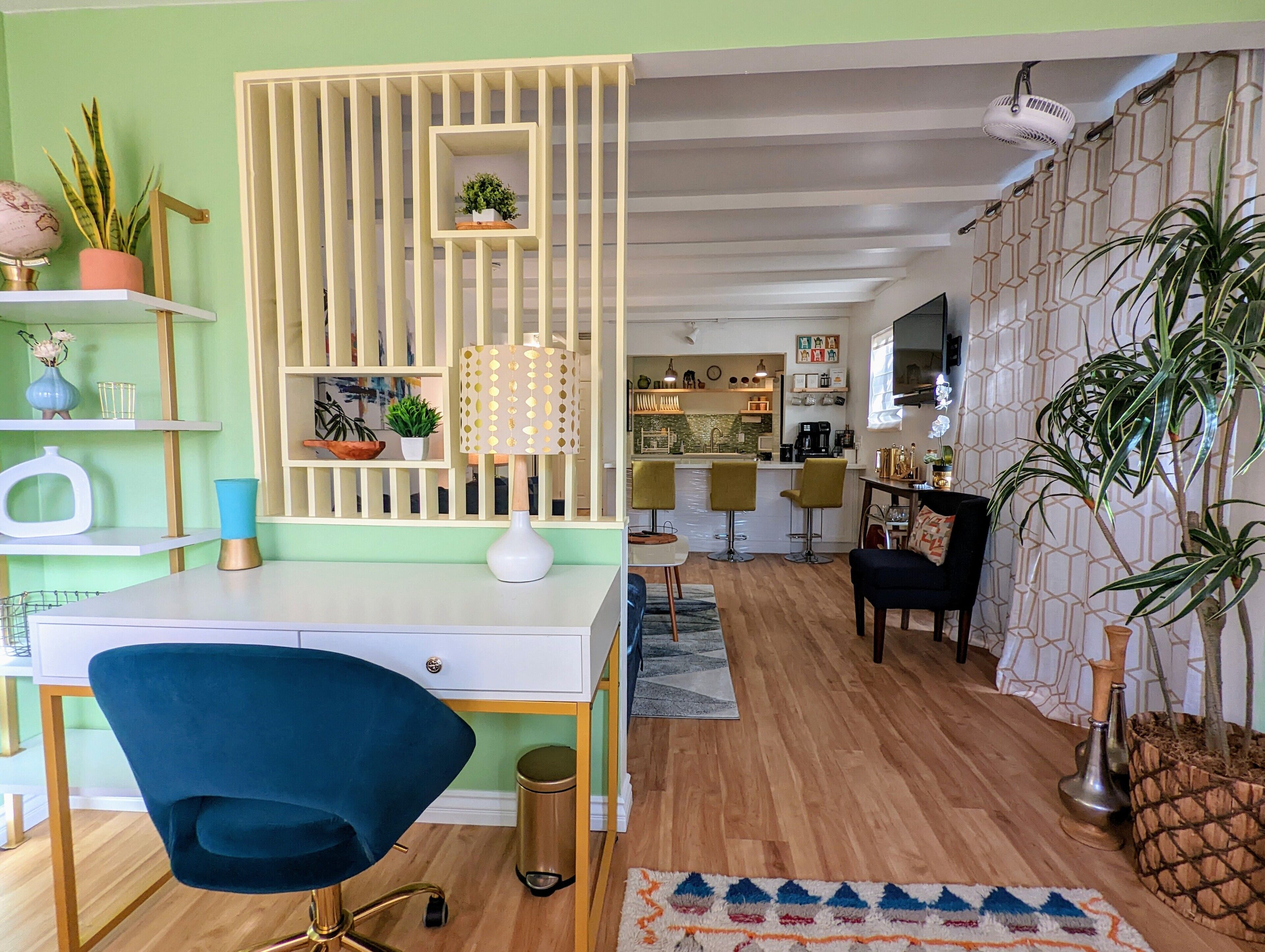 Un salon moderne avec un bureau, un fauteuil bleu, un élément de séparation décoratif et une cuisine en arrière-plan.
