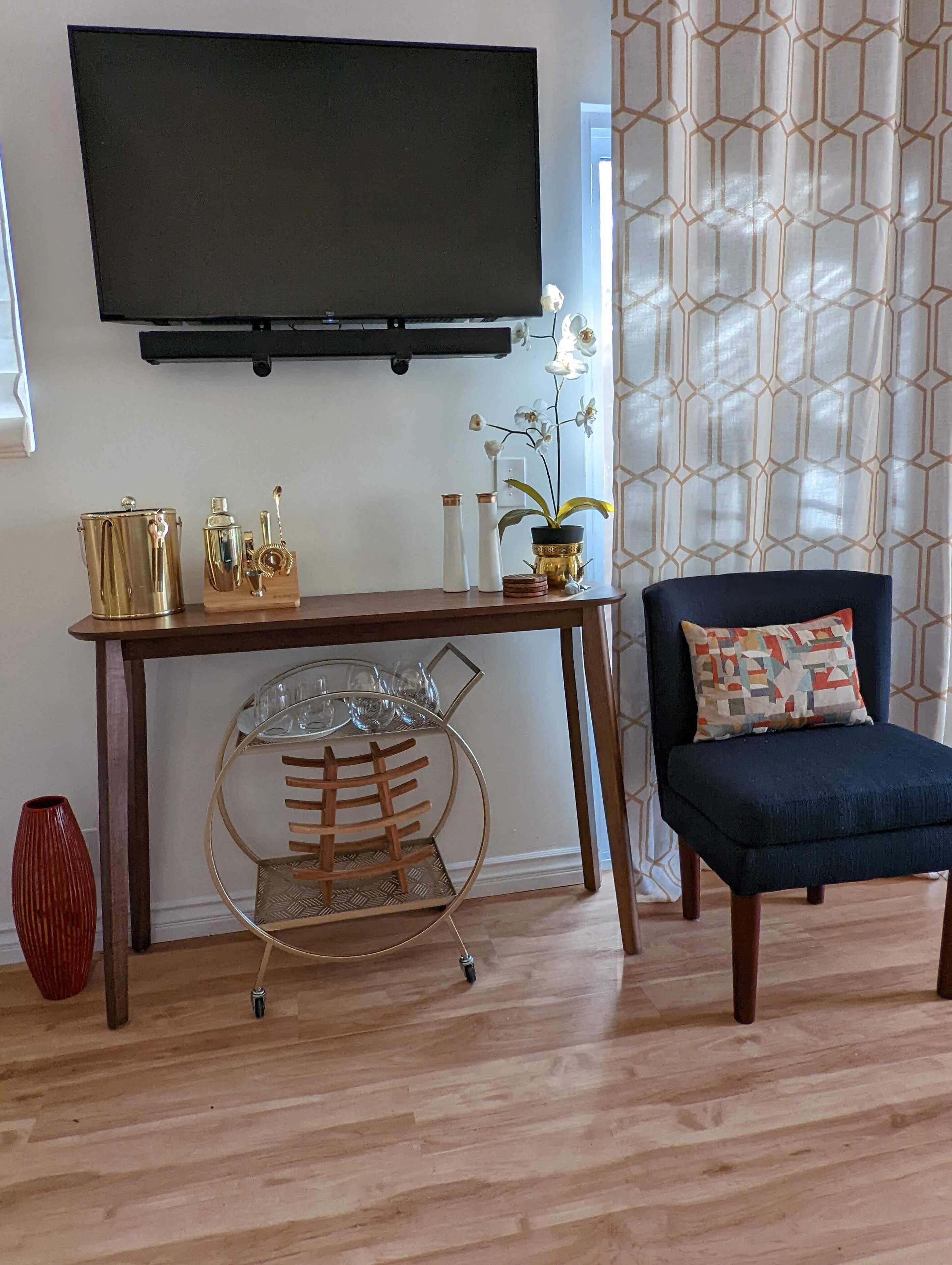 A living room with a TV mounted on the wall, a coffee table with decorative items, a chair with a patterned cushion, and a patterned curtain.
