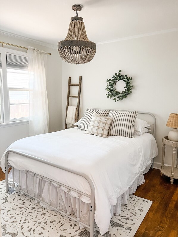 A bedroom with a white bed, a wooden ladder, a wicker side table, and a hanging light fixture.