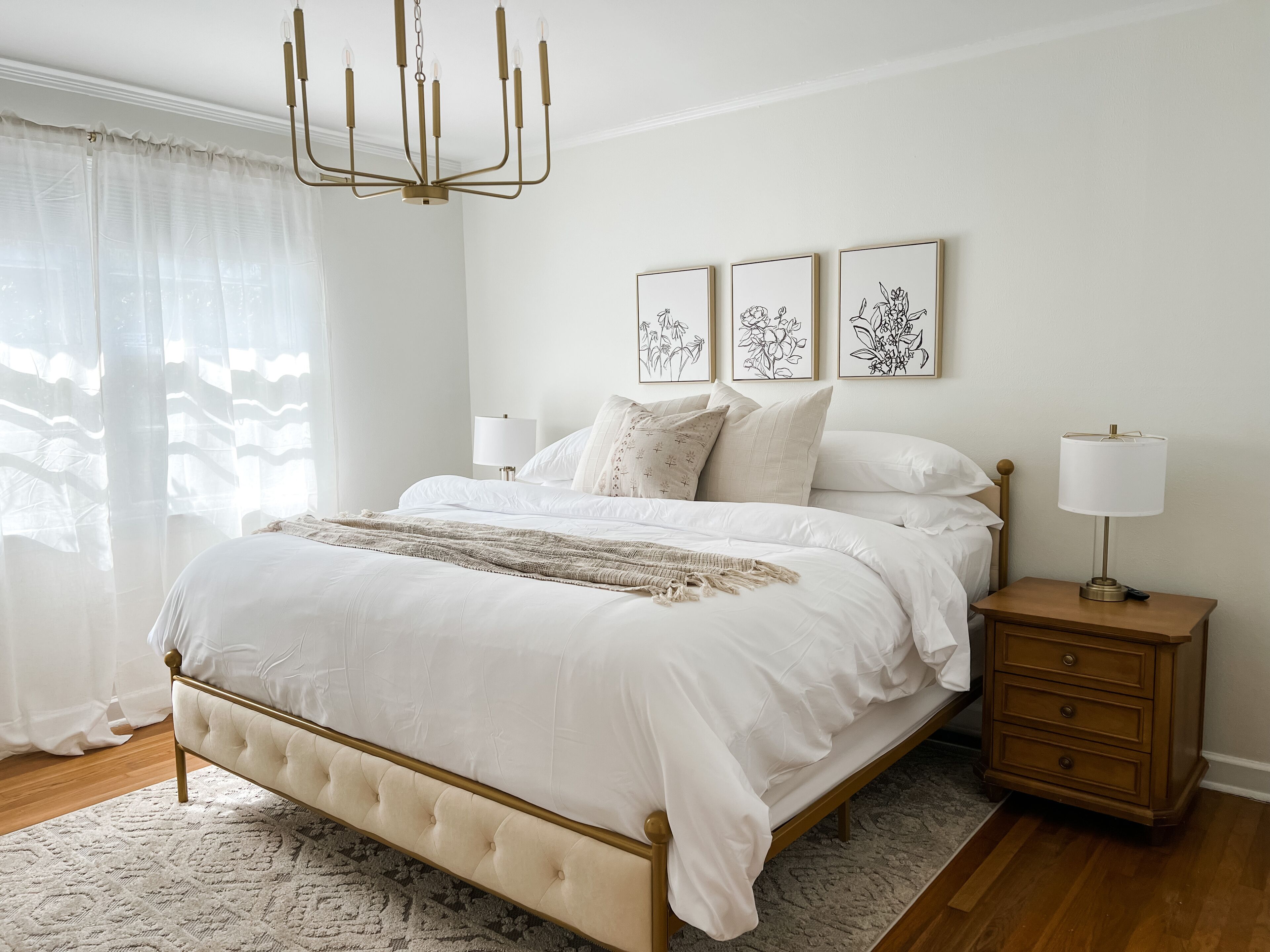 A bedroom with a large bed, a bedside table with a lamp, and three framed botanical prints on the wall.